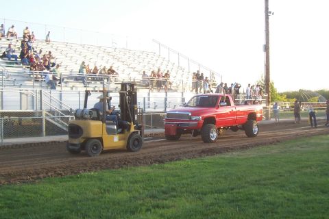 1994 Ford F-150 - A red pickup truck participates in a pull event, assisted by a forklift on a dirt track with spectators in the background.