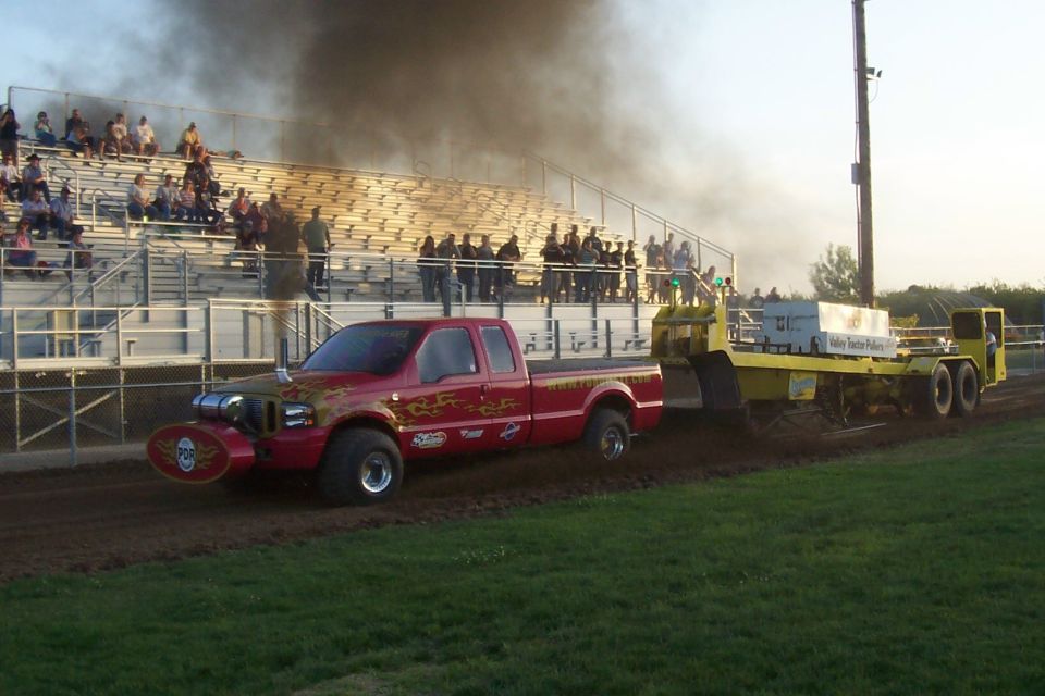 Red pickup truck with flames and skull decal on front, pulling a sled with a lot of black smoke coming out. Spectators in bleachers