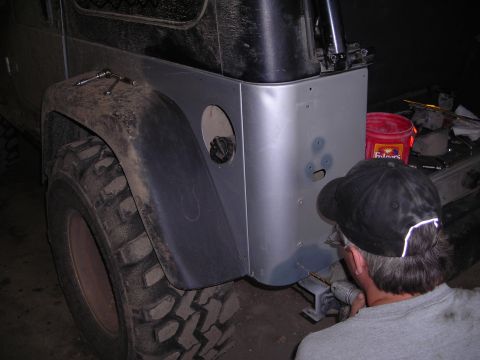 2003 Jeep TJ - A person drills into the rear quarter panel of a Jeep, with a large off-road tire in the foreground.