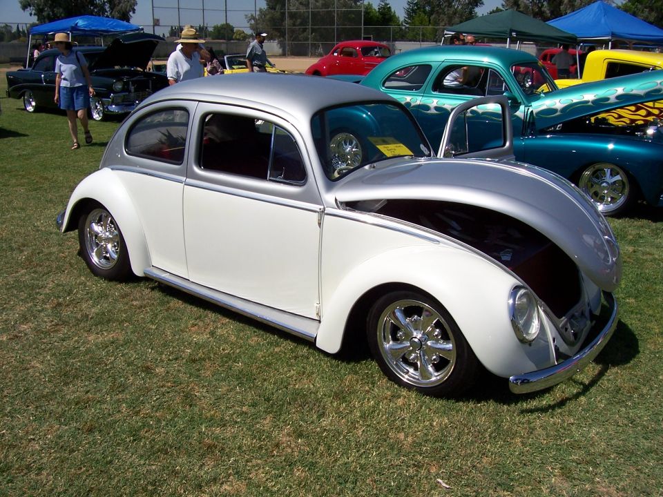Two-tone white and silver custom Volkswagen Beetle with chrome wheels, hood open. Other classic cars visible in background