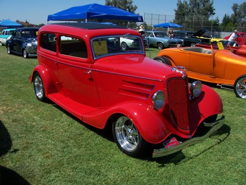 2002 Ford Mustang - A classic red hot rod sedan is parked on a grassy field at an outdoor car show.
