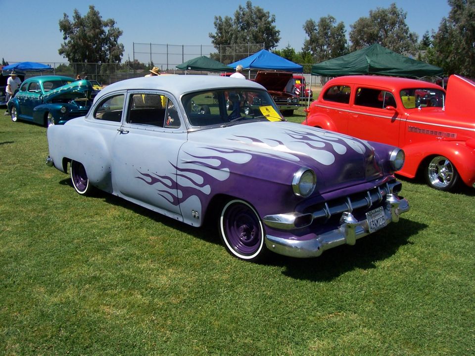 Light blue and purple classic car with flame graphics, parked on grass next to a red car