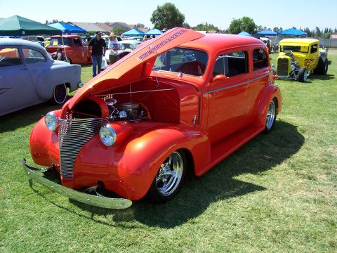 2002 Ford Mustang - A bright red hot rod sedan is displayed with its hood open at an outdoor car show.