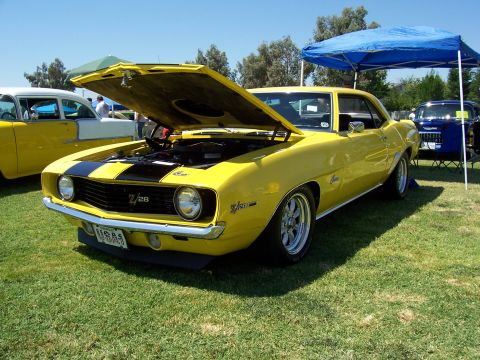 2002 Ford Mustang - A bright yellow 1969 Chevrolet Camaro Z/28 is displayed with its hood open, parked on a grassy field next to other vehicles.
