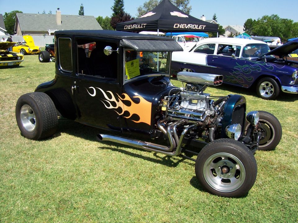 Black hot rod with orange flames, exposed engine, and custom wheels parked on grass