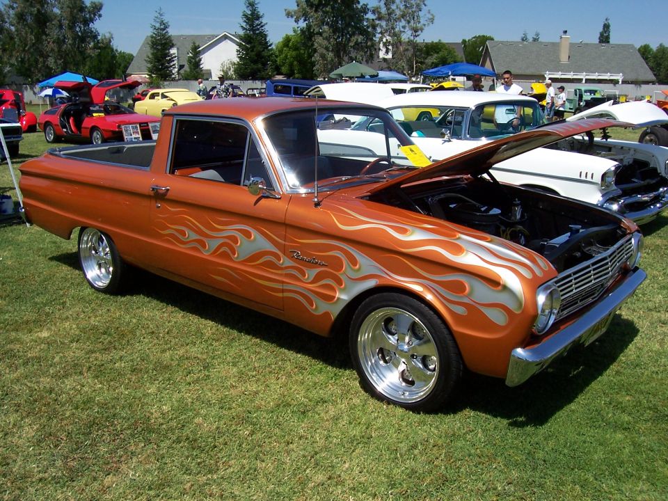 Orange 1965 Ford Falcon Ranchero pickup with silver flame decals, hood open, parked on grass at a car show