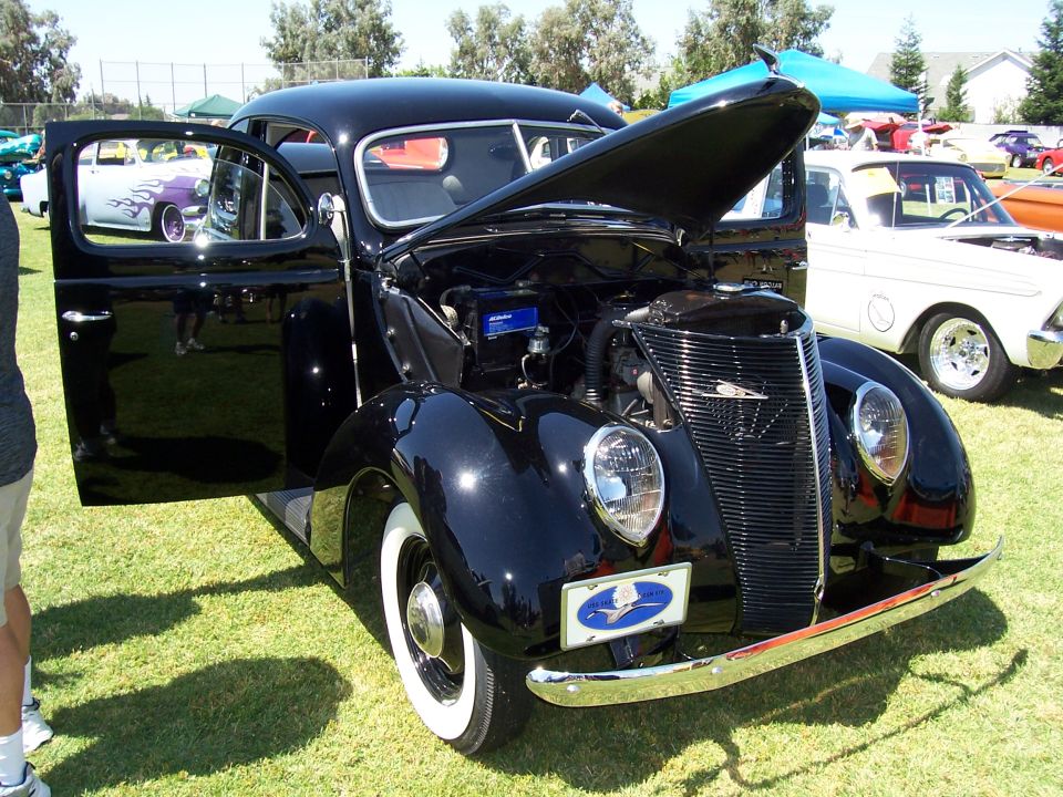 Black vintage car with hood and driver's door open, parked on grass with other cars in background