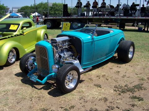 2002 Ford Mustang - A turquoise hot rod and a green custom pickup truck are parked outdoors at an event with a band on a trailer in the background.