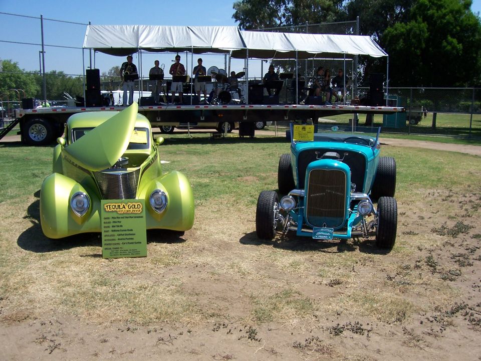 Green hot rod with hood up next to a blue hot rod. A band plays on a stage behind them