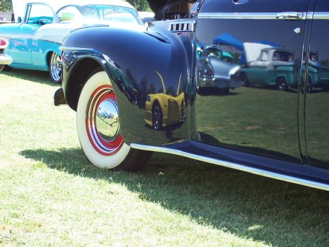 2002 Ford Mustang - Reflections on the polished surface of a dark vintage car reveal other vehicles and tents at an outdoor gathering.