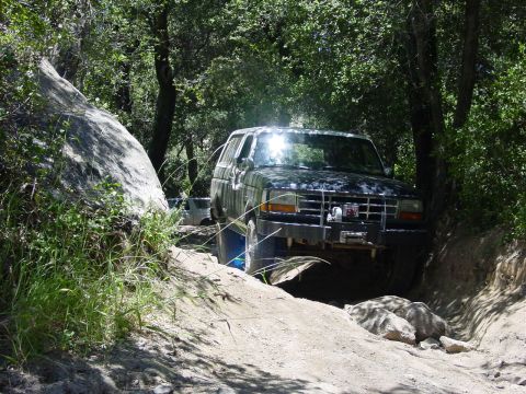 1994 Ford Bronco - A dark SUV navigates a rocky, wooded trail with another vehicle visible in the background.