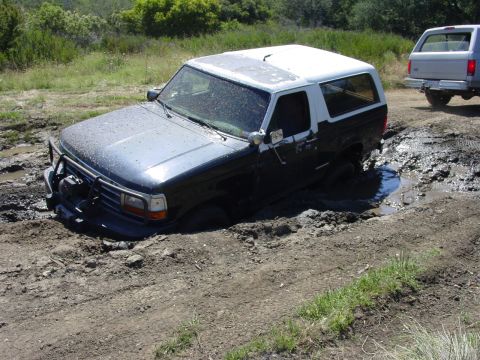 1994 Ford Bronco - A mud-spattered dark blue full-size Ford Bronco with a white hardtop is buried to the axles in a mud pit, front-left three-quarter view, we…