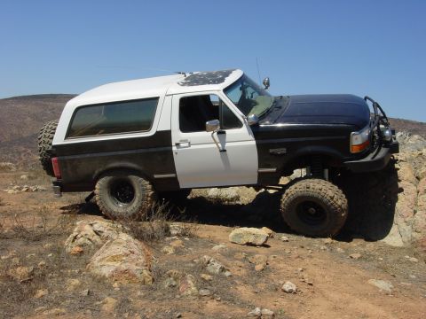 1994 Ford Bronco - Side profile of a lifted two-tone black and white Ford Bronco climbing a rocky dirt hillside under clear blue sky, on oversized mud-terrain…