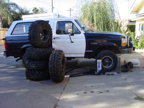 1994 Ford Bronco - A two-tone Ford Bronco is parked outside with a stack of tires and various suspension parts for what appears to be an upgrade.