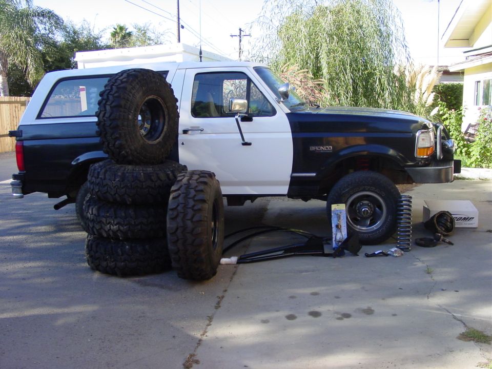 Two-tone Ford Bronco with stack of off-road tires and suspension parts laid out on the ground