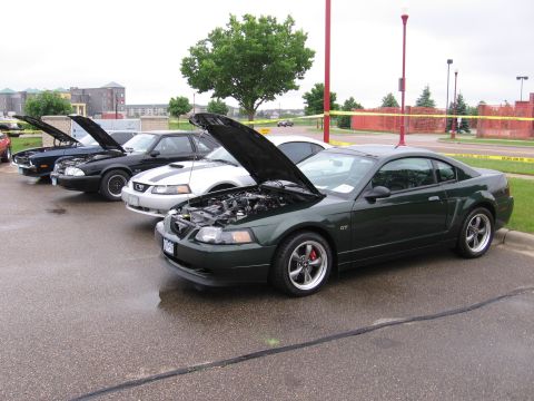 1989 Ford Mustang - A dark green SN95/New Edge Ford Mustang GT coupe with its hood raised sits at the end of a row of Mustangs in a wet parking lot at a car sh…