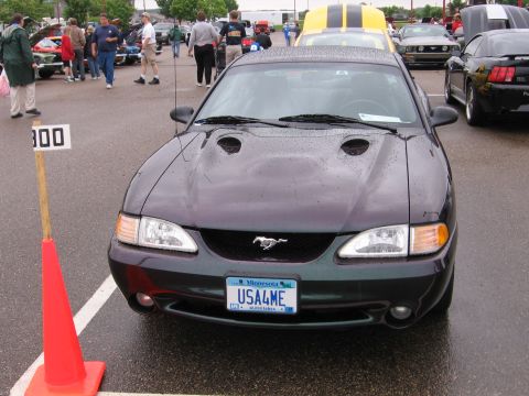 1989 Ford Mustang - A dark colored Ford Mustang is parked at an outdoor car show with other vehicles and spectators present.