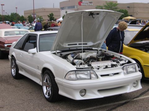1989 Ford Mustang - White Ford Mustang at a car show, hood open to display the engine. Other vehicles and people are visible in the background.