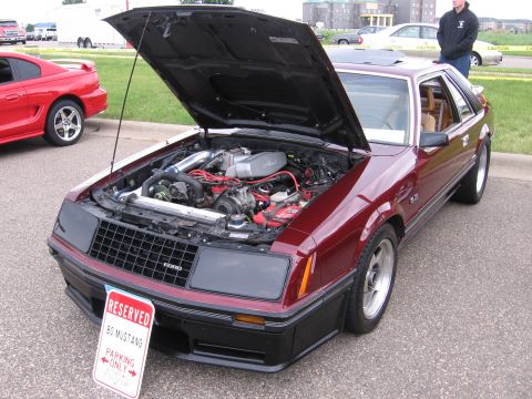 1989 Ford Mustang - A maroon Ford Mustang is parked with its hood up, revealing the engine bay, while another red Mustang is visible in the background.