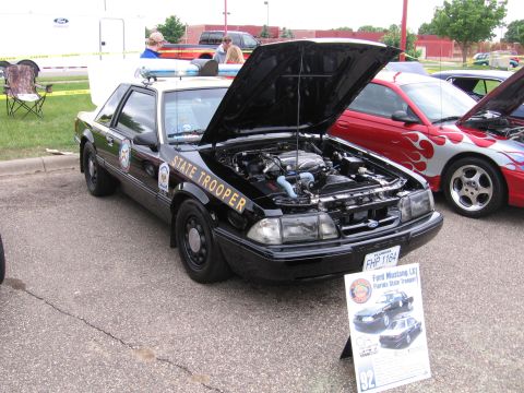 1989 Ford Mustang - A black Ford Mustang police car with its hood open is displayed next to a red car with flames.