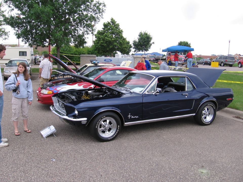 Dark blue classic car with flame decals, hood open, parked at a car show. Other cars and people are visible