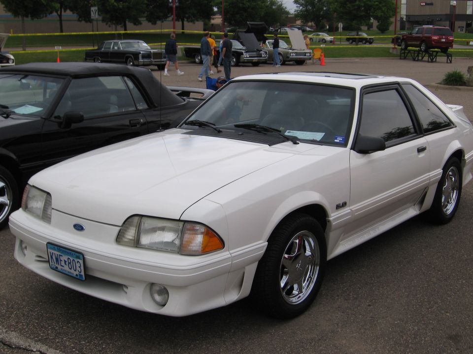 White 1989 Ford Mustang coupe with chrome wheels. Black convertible parked next to it. Other cars and people in background at a car show