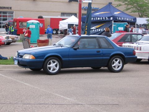 1989 Ford Mustang - A blue coupe is parked at an outdoor event with tents and trailers in the background.