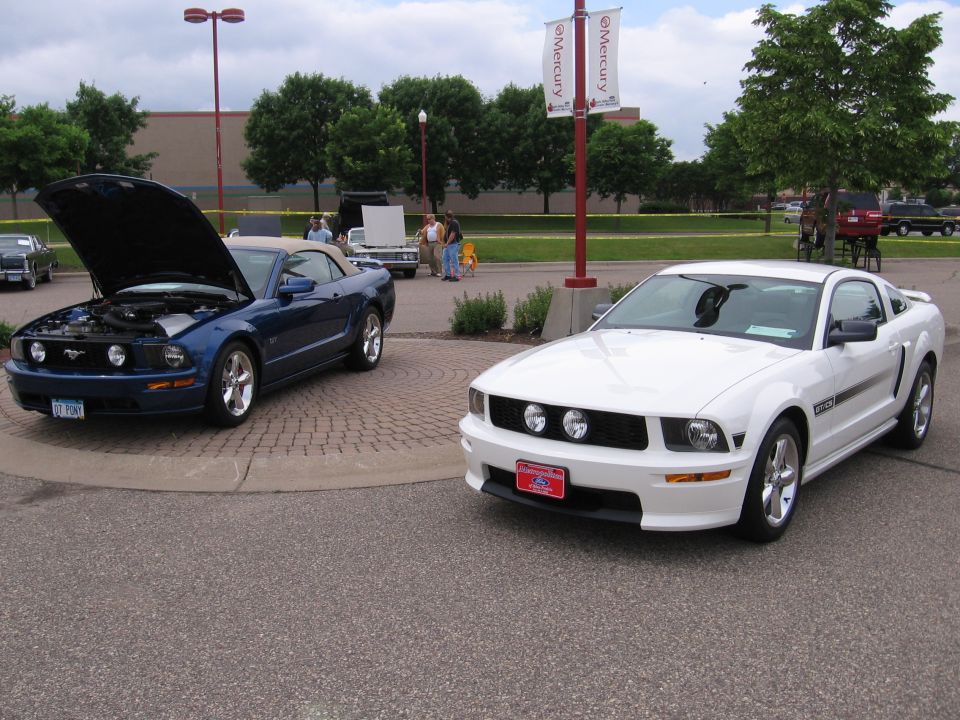 White Ford Mustang parked next to a blue convertible Mustang with its hood up