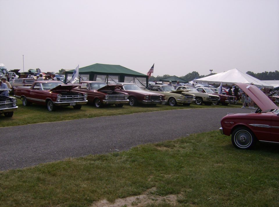 Several classic cars lined up at an outdoor event with tents in the background
