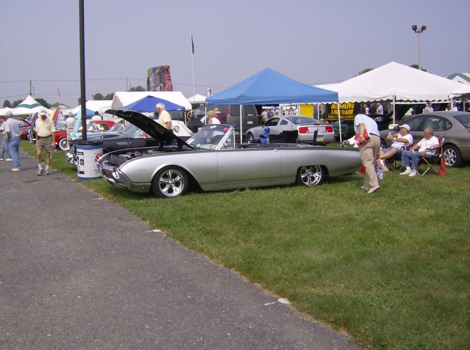 Silver convertible with hood up, parked on grass at an outdoor car show. Other cars and tents in background