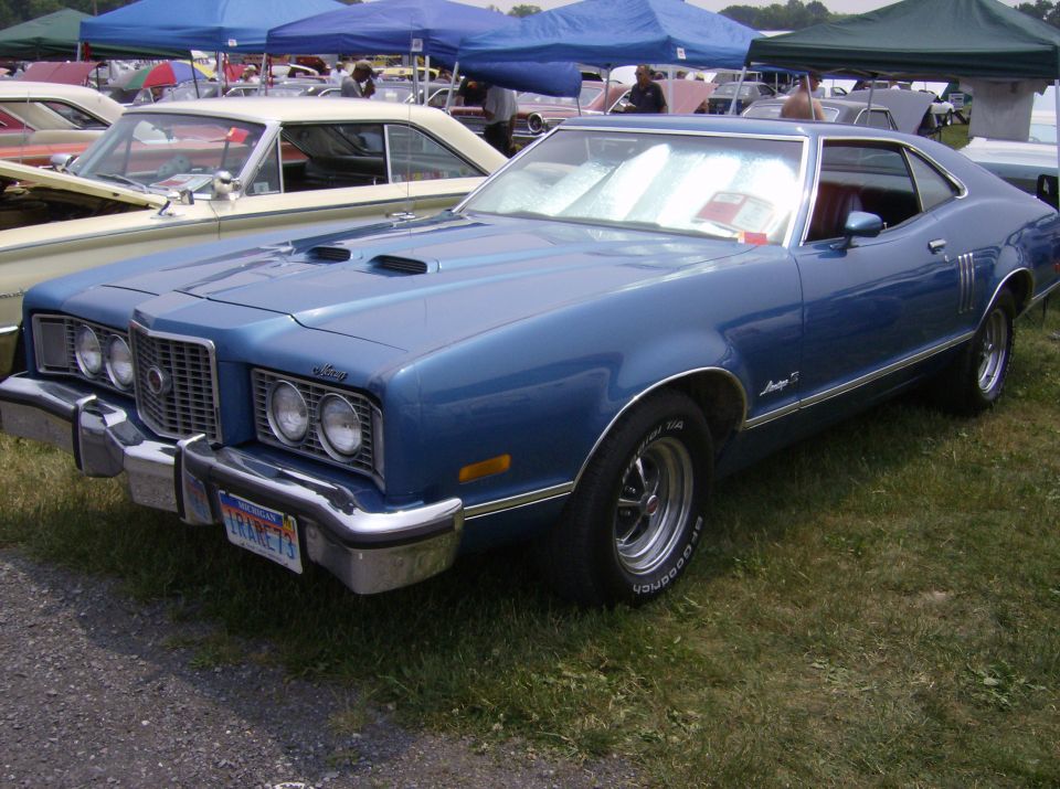 Blue coupe with hood scoops and chrome grille, parked on grass next to another car. License plate reads 'FRANKE73'