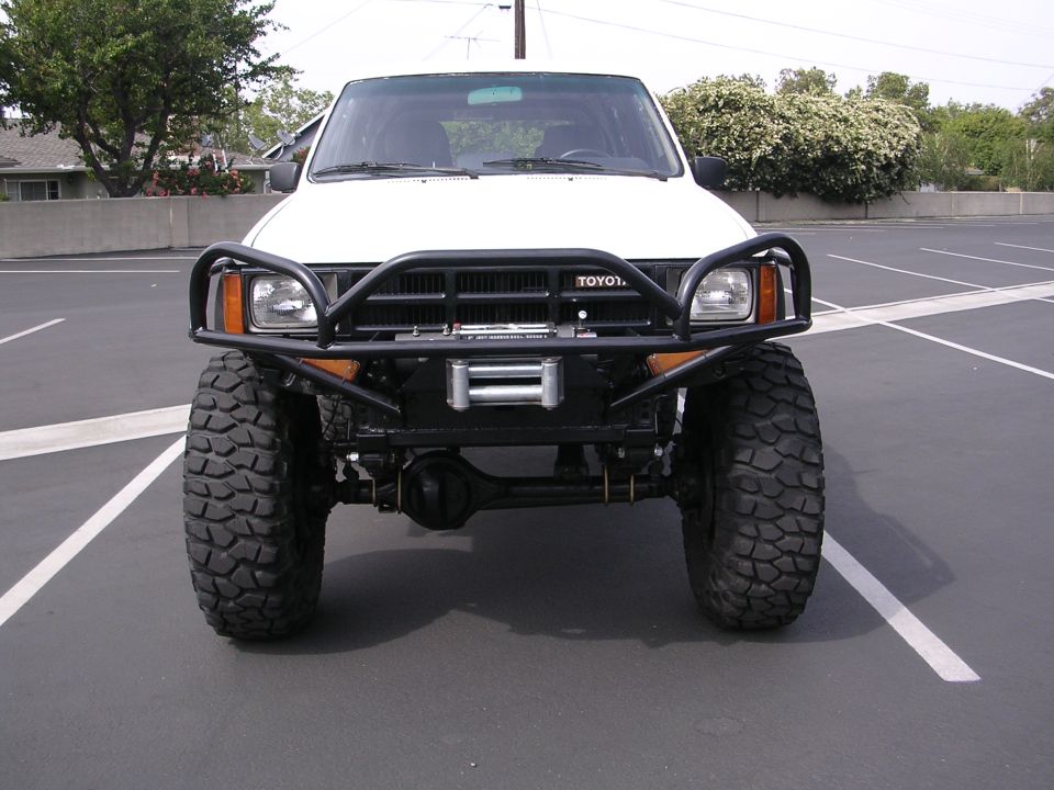 Front view of a white Toyota pickup truck with a black aftermarket bumper, winch, and large off-road tires. The grille has 'TOYOTA' text