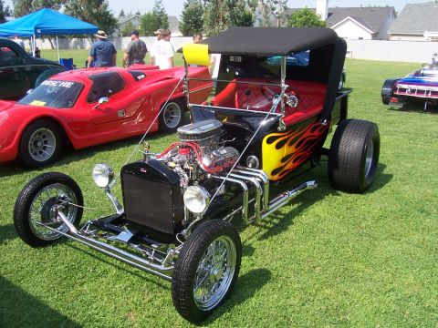 2002 Ford Mustang - A customized T-bucket hot rod is displayed on grass next to other vehicles at an outdoor event.