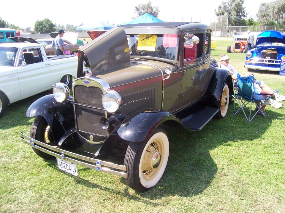 1930 Ford Model A coupe with hood open, parked on grass next to a white pickup truck