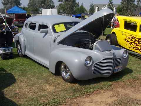 2002 Ford Mustang - A customized gray sedan is displayed with its hood open, alongside a yellow hot rod at an outdoor event.