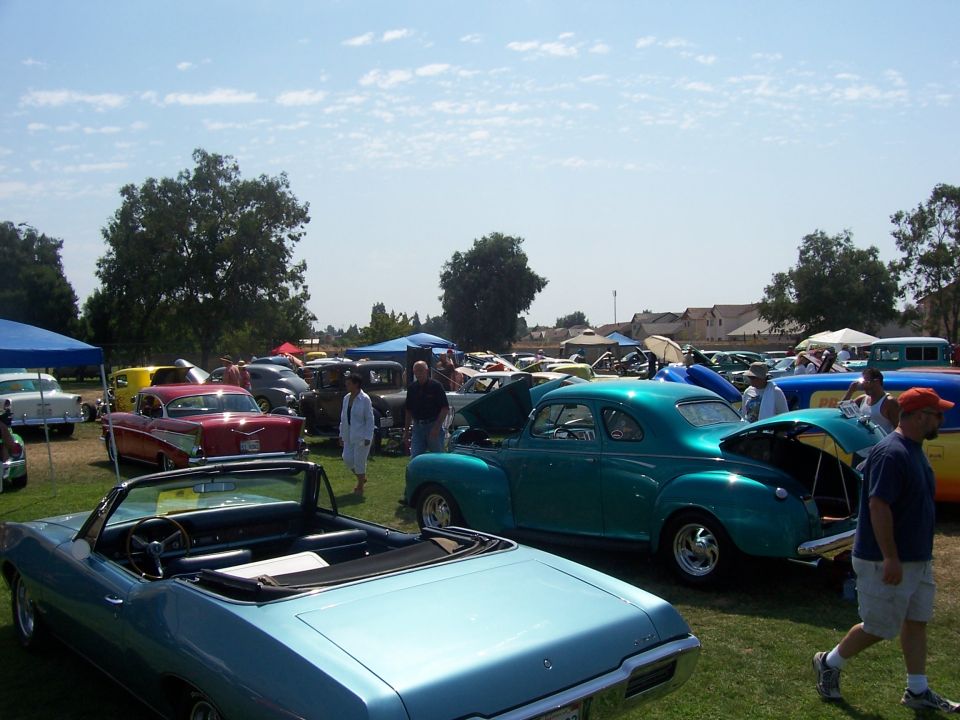 Teal coupe with open trunk and hood, blue convertible in foreground, at an outdoor car show