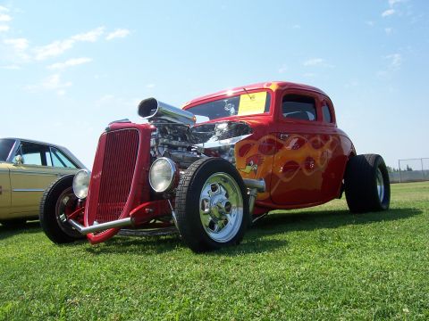 2002 Ford Mustang - A custom red hot rod with flame decals and a prominent engine sits on a grassy field next to another car.