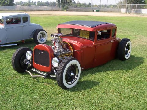 2002 Ford Mustang - A vintage red hot rod with a prominent engine sits on a grassy field, with another classic car visible behind it.