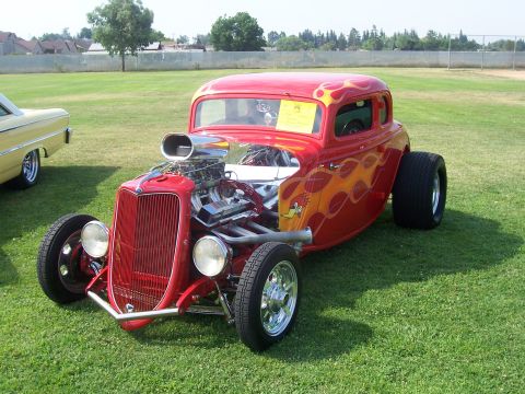 2002 Ford Mustang - A red hot rod with flame graphics and a prominent supercharger sits on a grassy field next to another car.