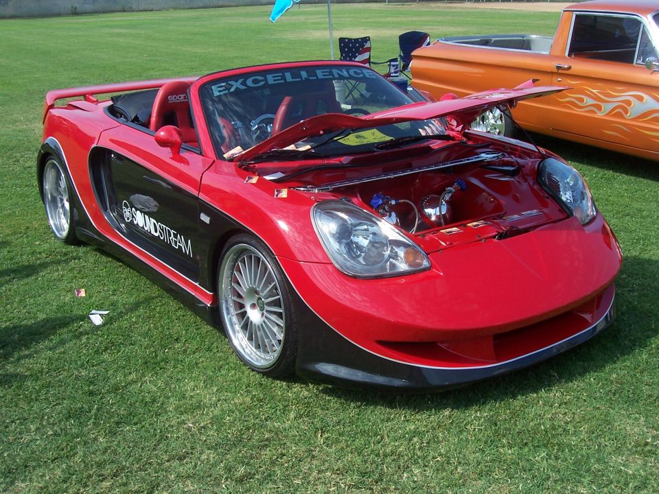 Red and black convertible sports car with hood open, parked on grass next to an orange pickup truck