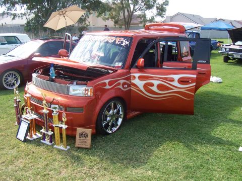 2002 Ford Mustang - An orange hatchback with custom flame decals and open doors is displayed with trophies.