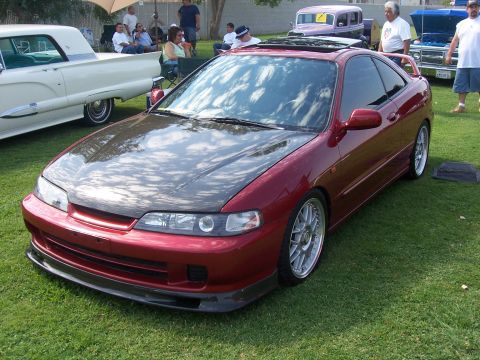 2002 Ford Mustang - A red coupe with a carbon fiber hood and aftermarket wheels is displayed on a grassy lawn, with other vehicles and people in the background.