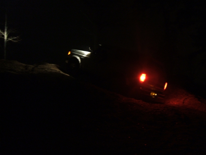 SUV driving on a dirt path at night, headlights on, taillights glowing red