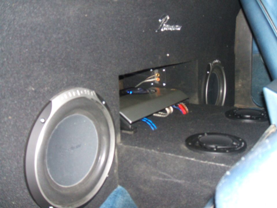 Car interior showing a large subwoofer, an amplifier, and two smaller speakers mounted in the carpeted rear area