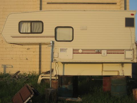 1996 Dodge 2500 - A side view of a truck camper parked in front of a brick building, resting on makeshift supports.