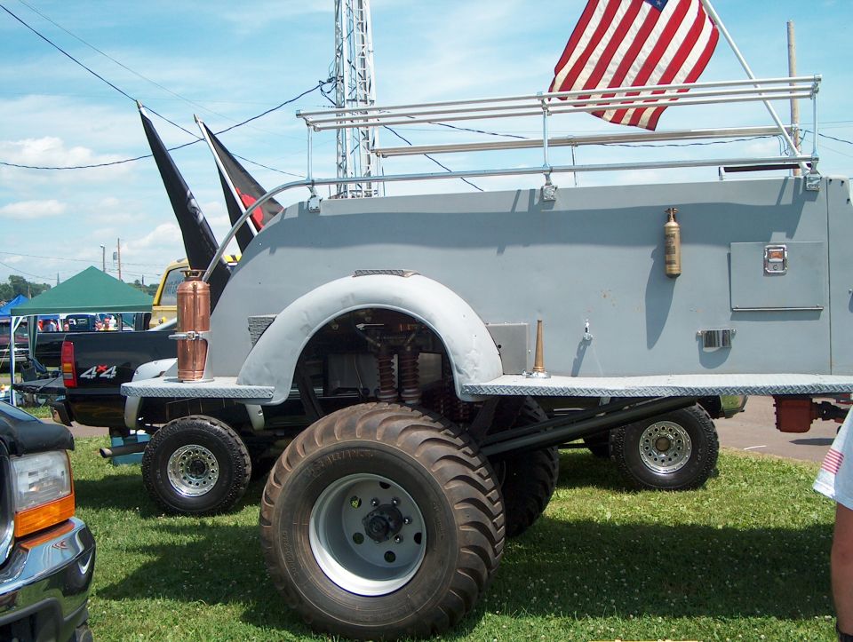 Custom truck with large tires, American flag on roof rack, and other vehicles in background