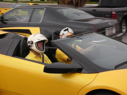 1998 Ford Crown Victoria - Two individuals in helmets are driving a yellow convertible, with other vehicles visible in the background.