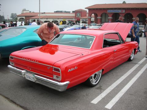 1998 Ford Crown Victoria - A red 1965 Chevrolet Malibu SS is parked at a car meet, with its hood open and people gathered around.