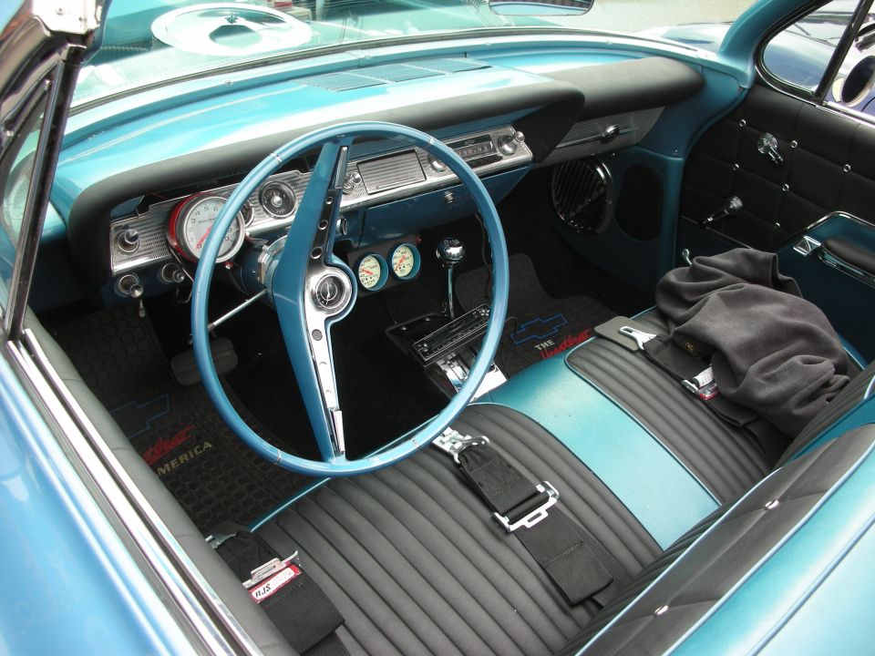 Blue interior of a classic car with a large steering wheel, dashboard gauges, and bucket seats. Floor mats show a Chevrolet logo and 'The Impala'