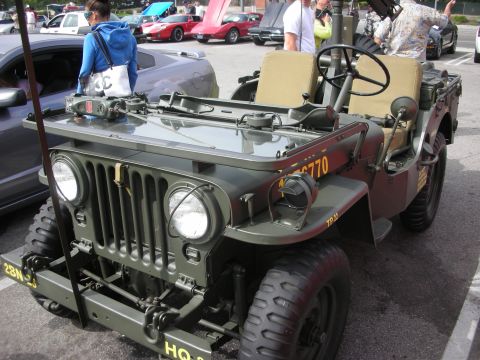 1998 Ford Crown Victoria - An olive drab military jeep is parked in a lot with other vehicles and people around.